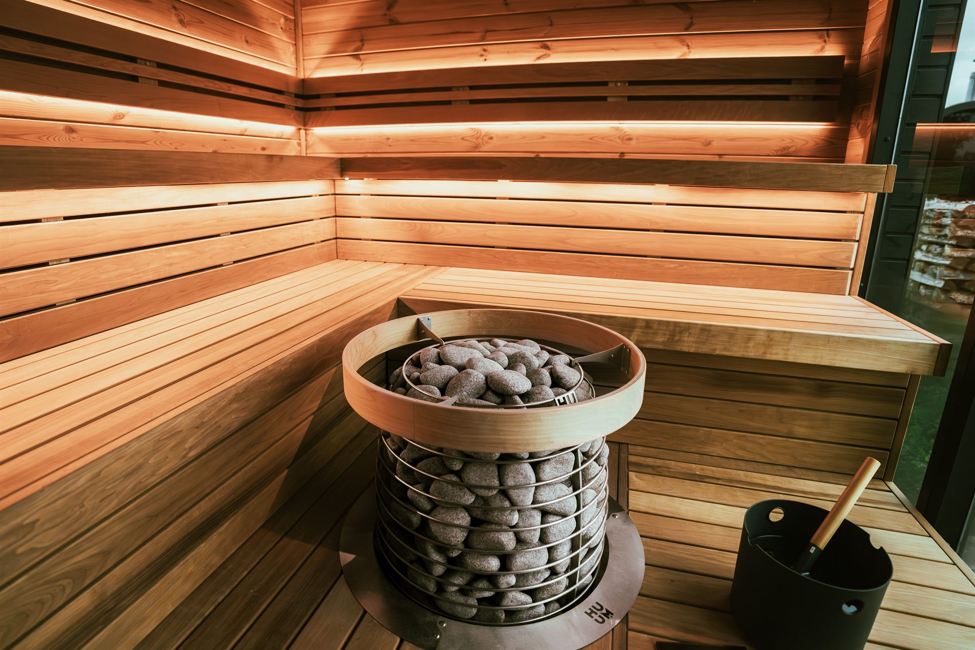 Sauna interior corner with cedar benches, a stone heater and a water bucket with ladle
