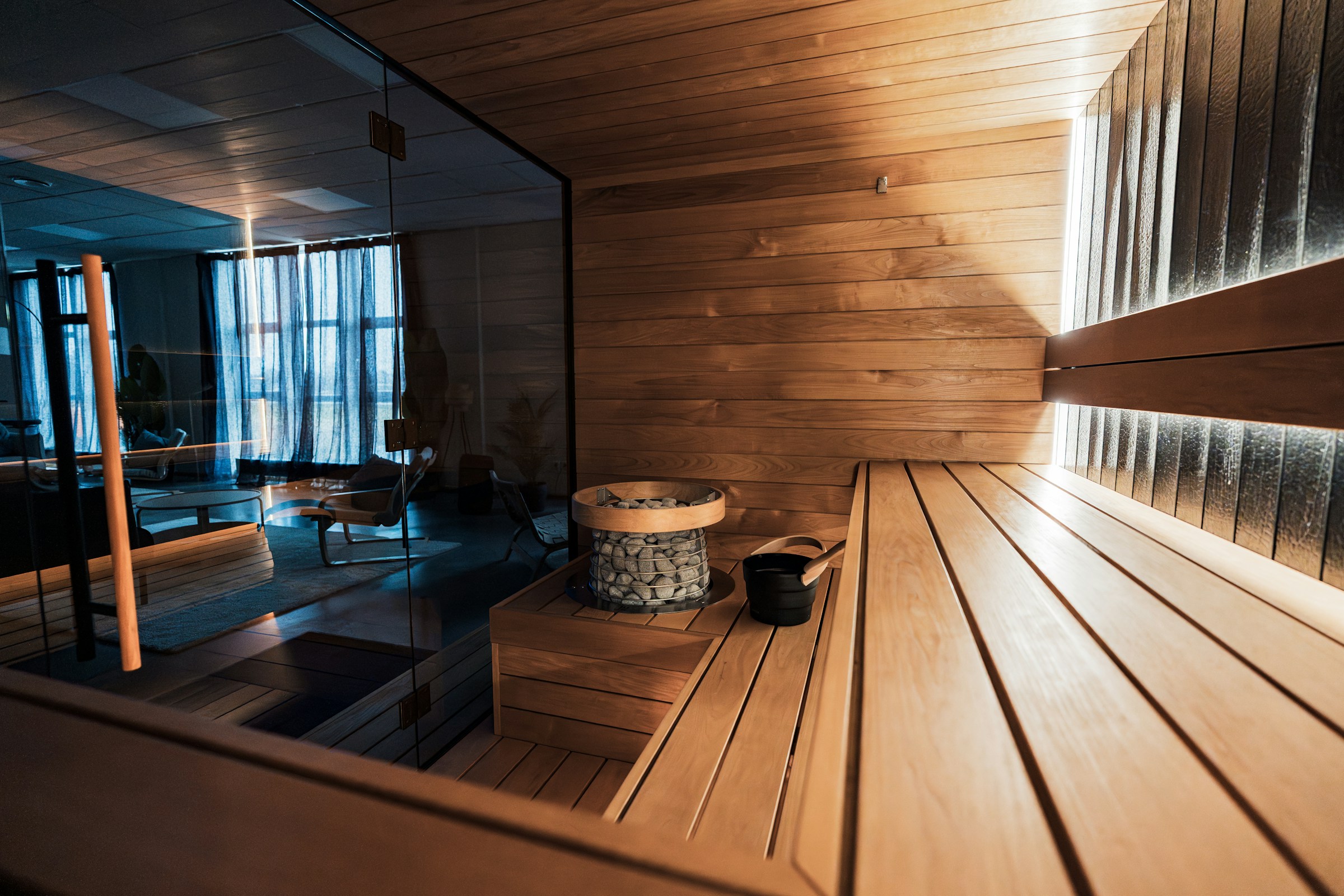 Sauna bench and stone heater beside a glass wall, with a lounge area visible beyond
